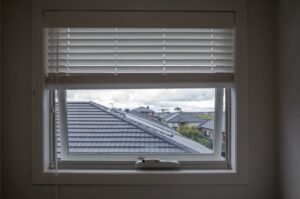 Awning window with white trim and blinds, overlooking a neighborhood roofline. Bow